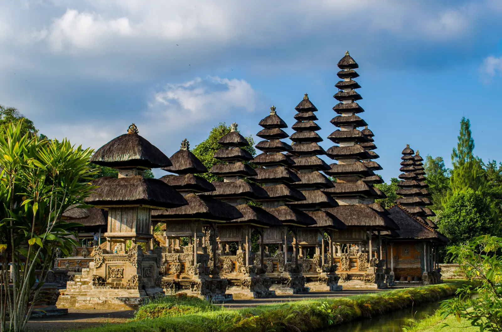A row of tall, black-thatched tiered shrines inside Pura Taman Ayun under a cloudy blue sky, with a canal in front
