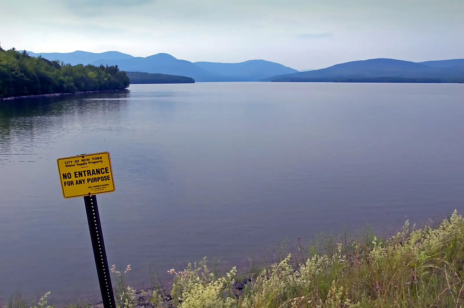 The calm water of the Ashokan Reservoir with blue Catskill ridges beyond and a yellow New York City water-supply sign on the bank
