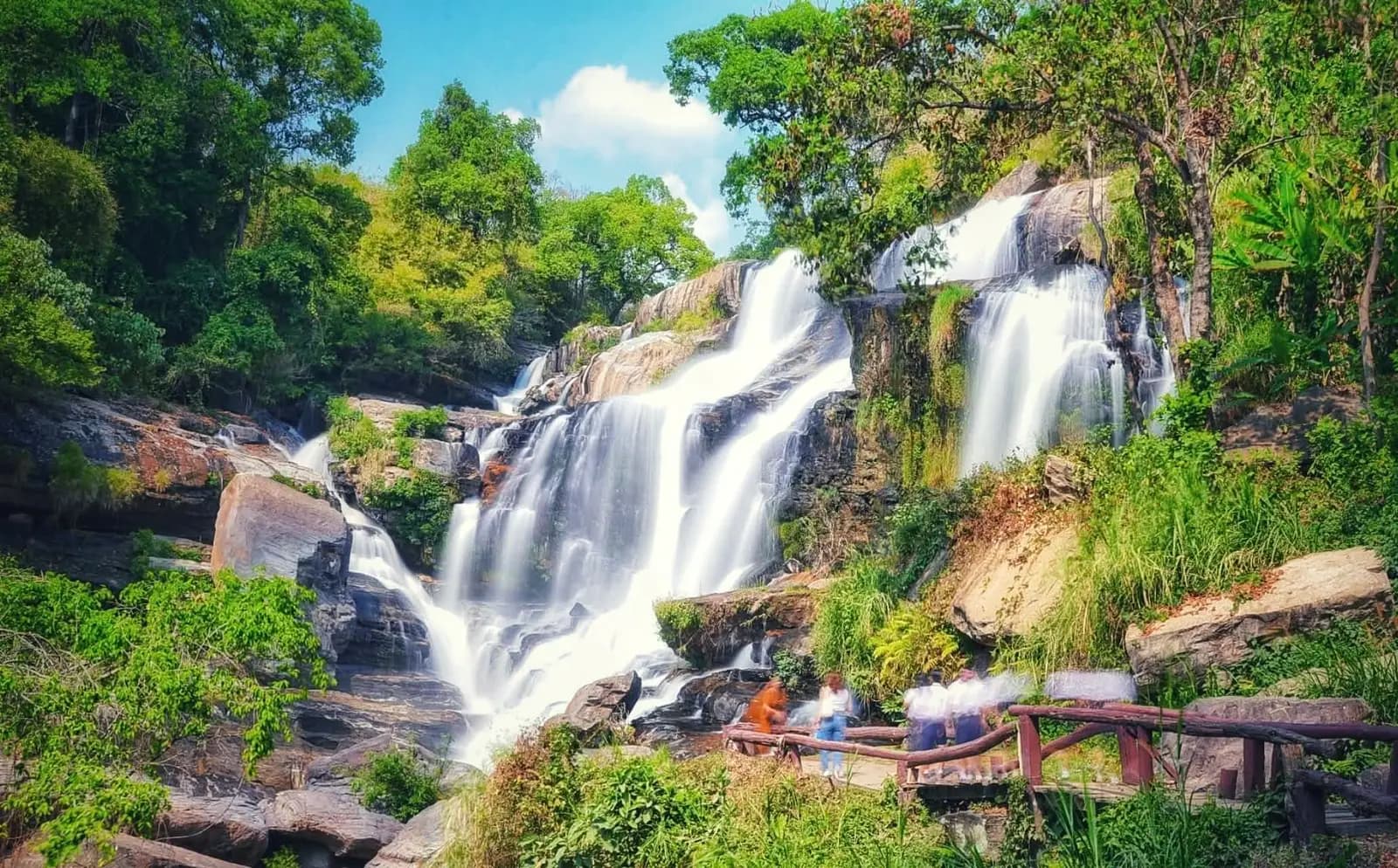Mae Klang Waterfall spilling over rock ledges among trees, with a wooden railing in the foreground