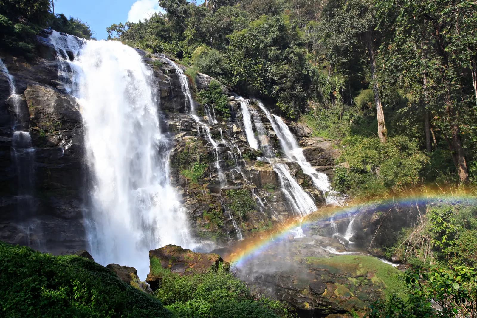 Wachirathan Waterfall pouring down a dark rock face, with a rainbow in its spray