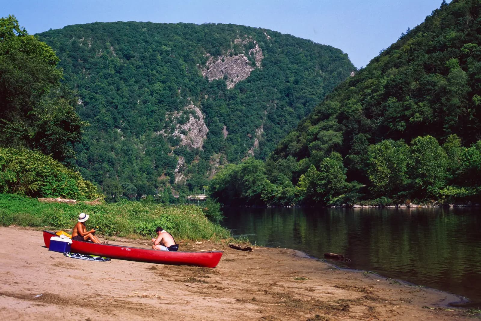 A red canoe on a sandy river beach below the steep forested cliffs of the Delaware Water Gap