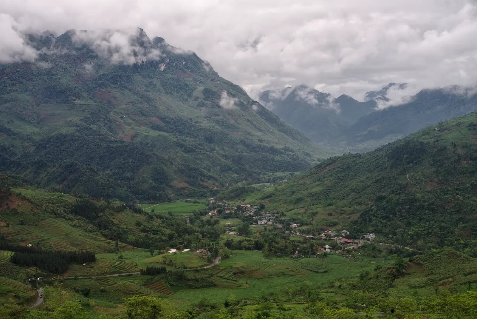 A green valley with a small village and terraced fields beneath cloud-capped mountains, seen from the Bac Sum climb