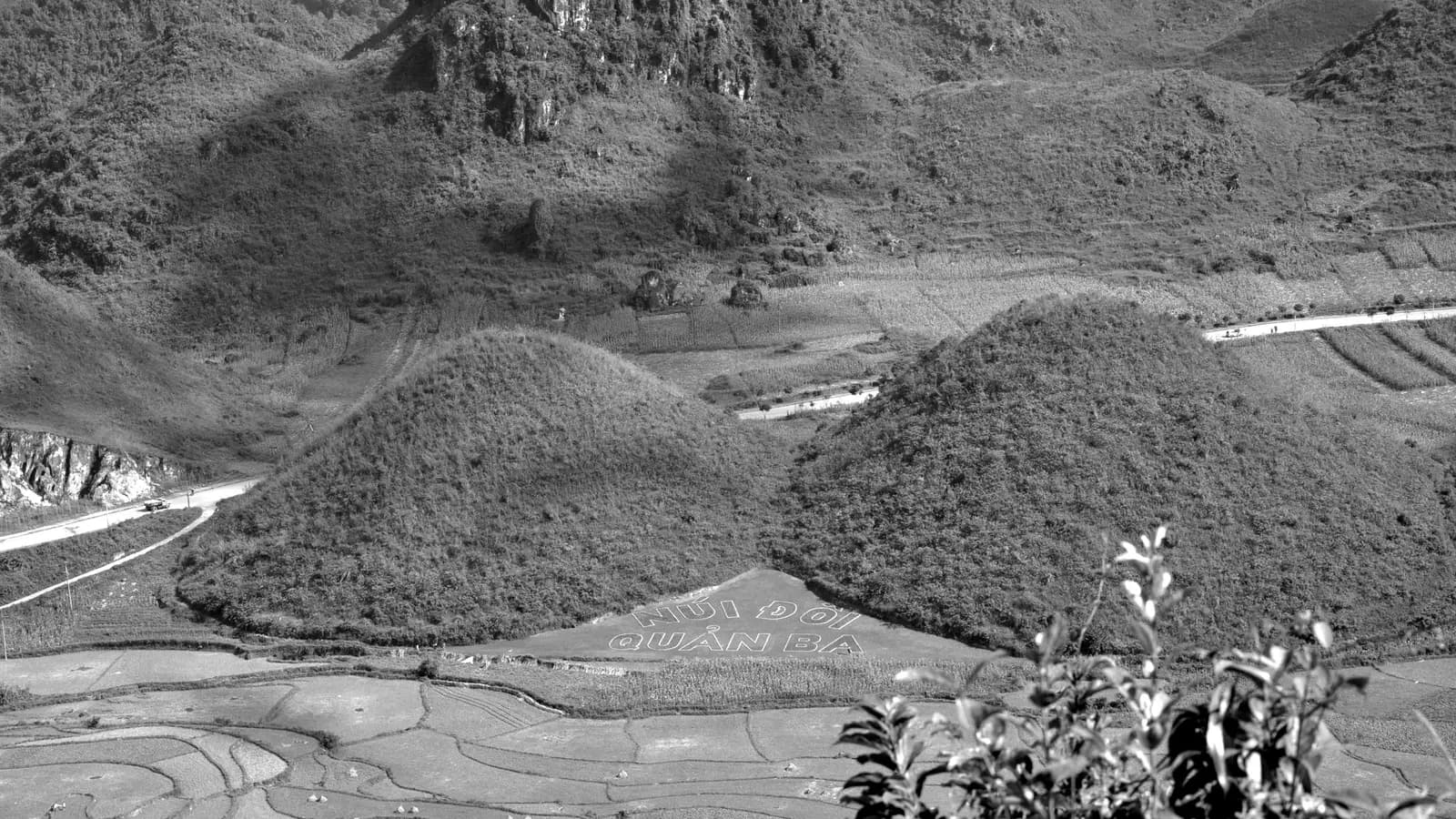 Black-and-white photo of the two rounded twin mountains of Quan Ba above paddy fields, with the road winding behind them