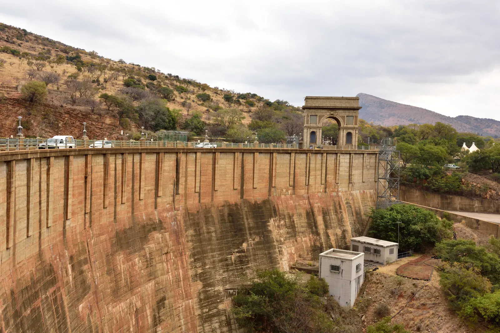 The curved concrete wall of the Hartbeespoort Dam with the road along its top and the stone arch at the far end