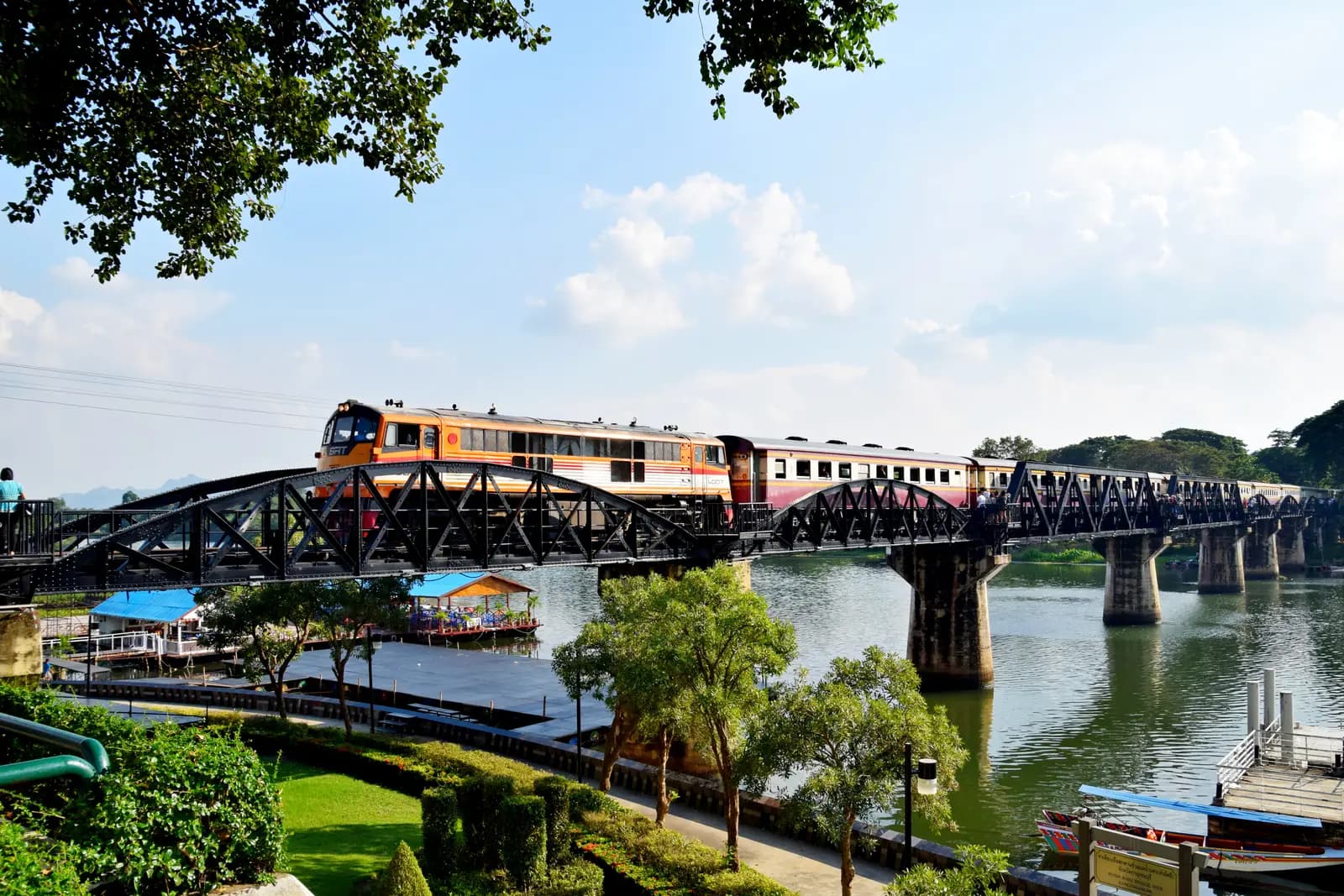A diesel passenger train crossing the Bridge on the River Kwai above the river, seen from the riverside garden