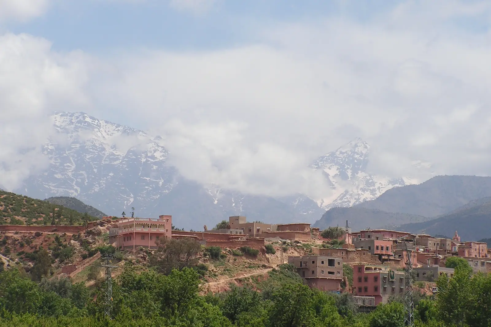 Pink houses of Asni on a hillside, with snow-streaked High Atlas peaks in cloud behind