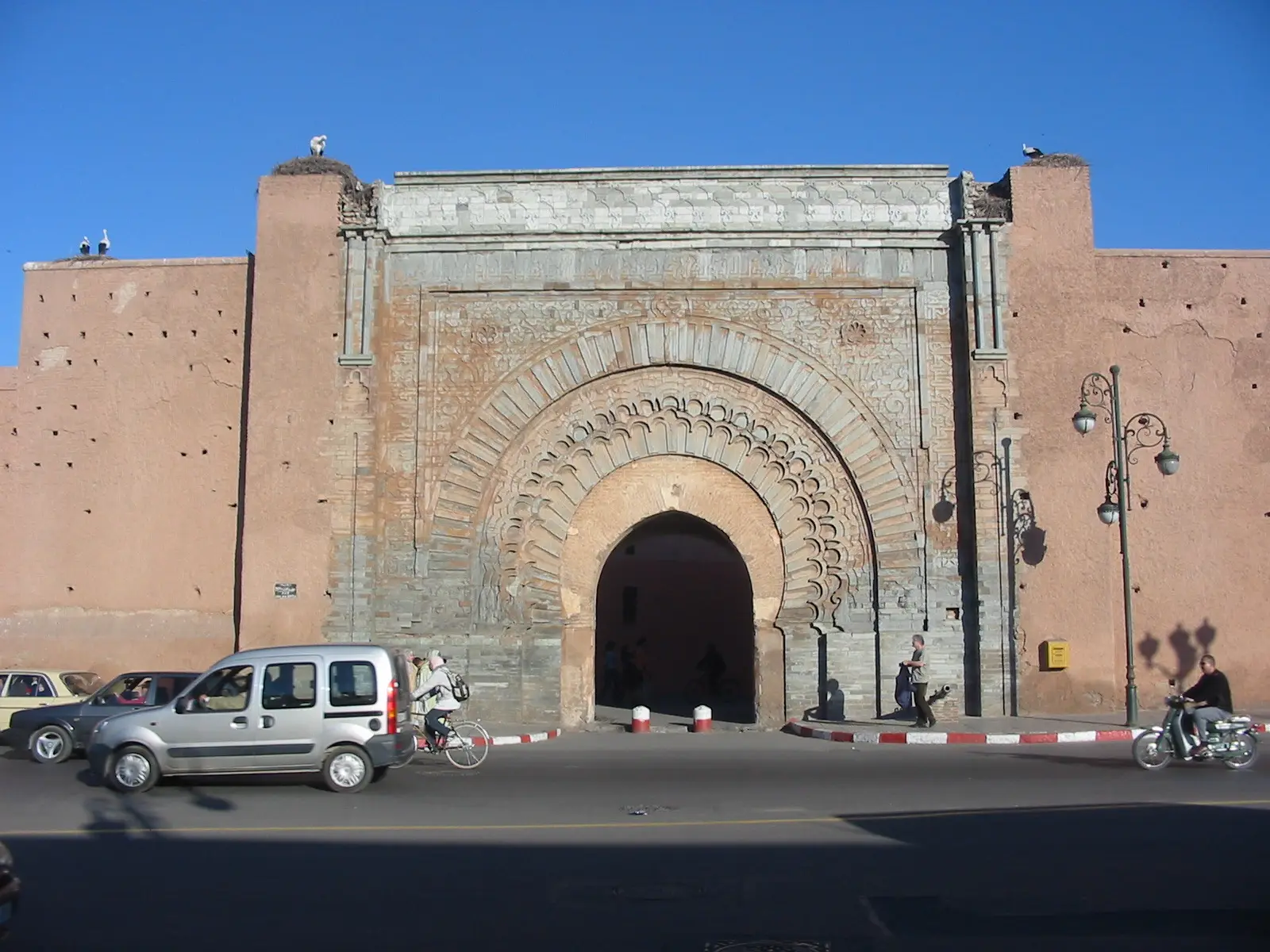 The carved stone arch of Bab Agnaou set in the red Kasbah wall, with a car and scooters passing