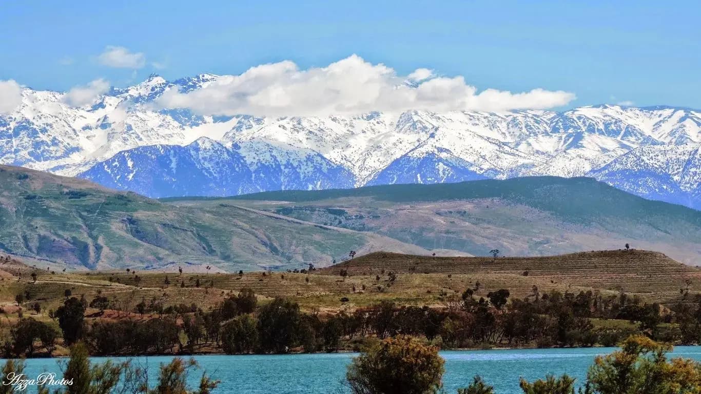 The blue water of Lalla Takerkoust reservoir below green hills, with the snow-covered High Atlas behind