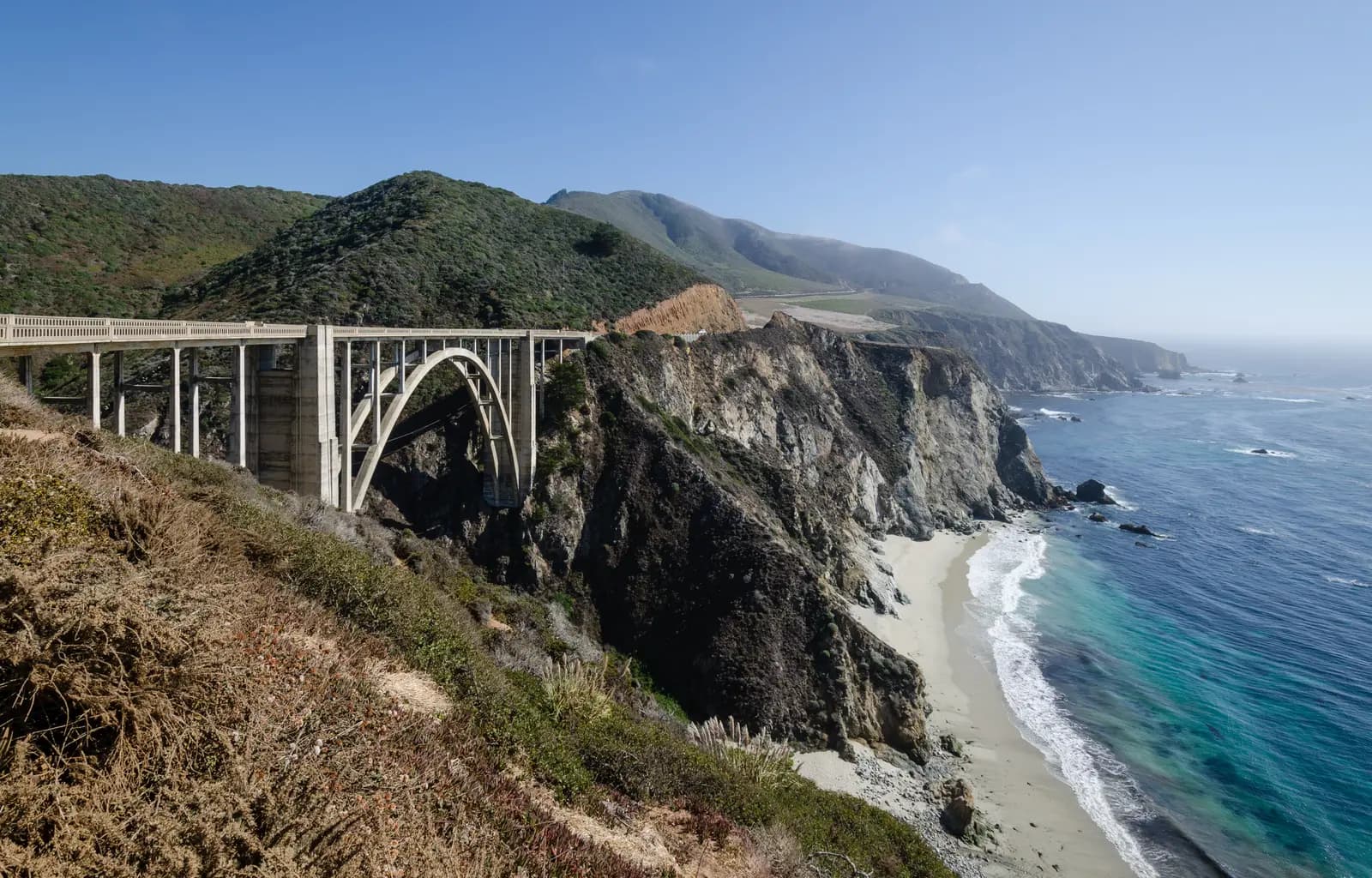 The concrete arch of Bixby Creek Bridge above a sandy cove, with the coast running south