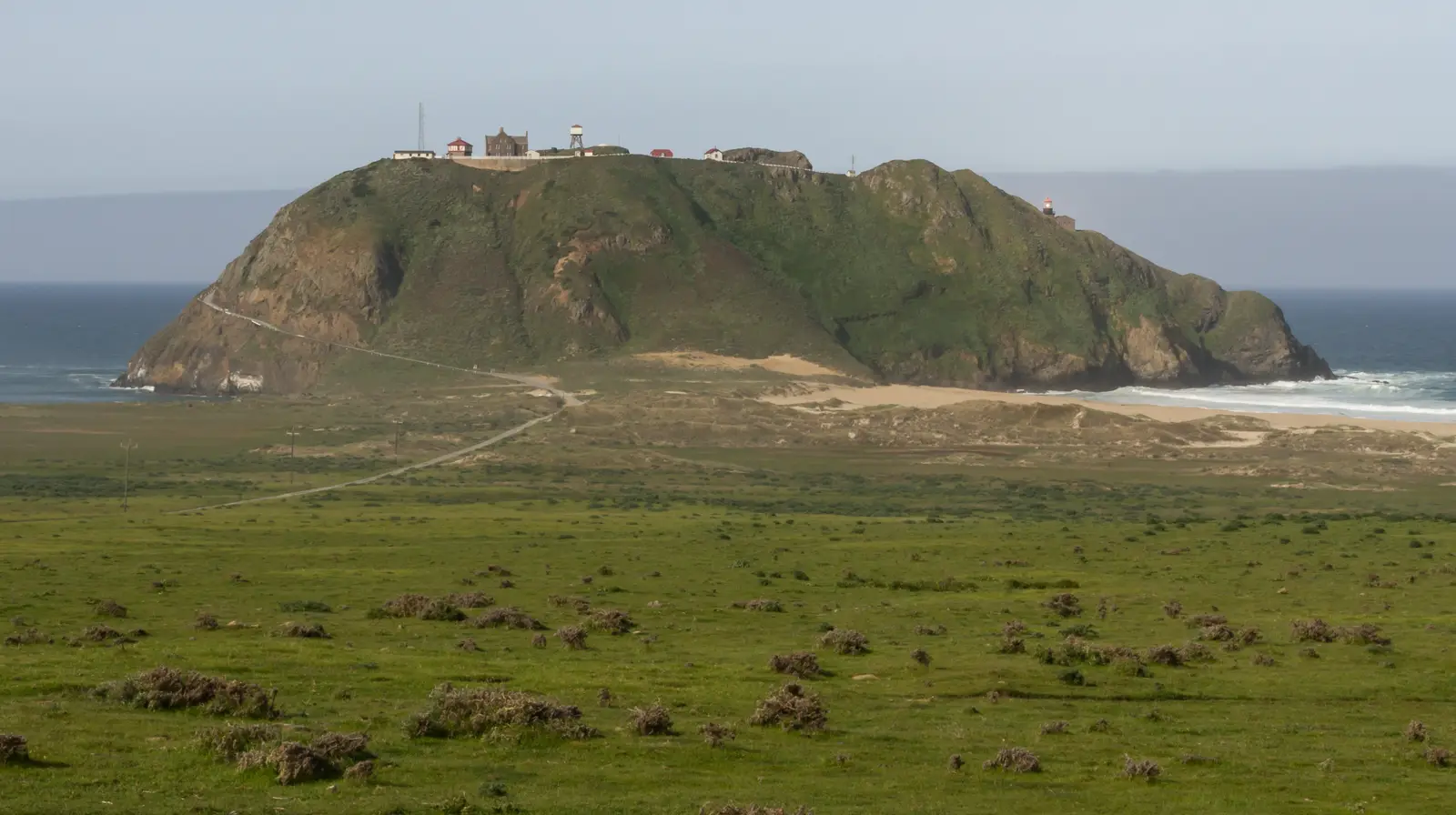 Point Sur, a green rock with the light station buildings on top, rising above pasture and a sandy beach