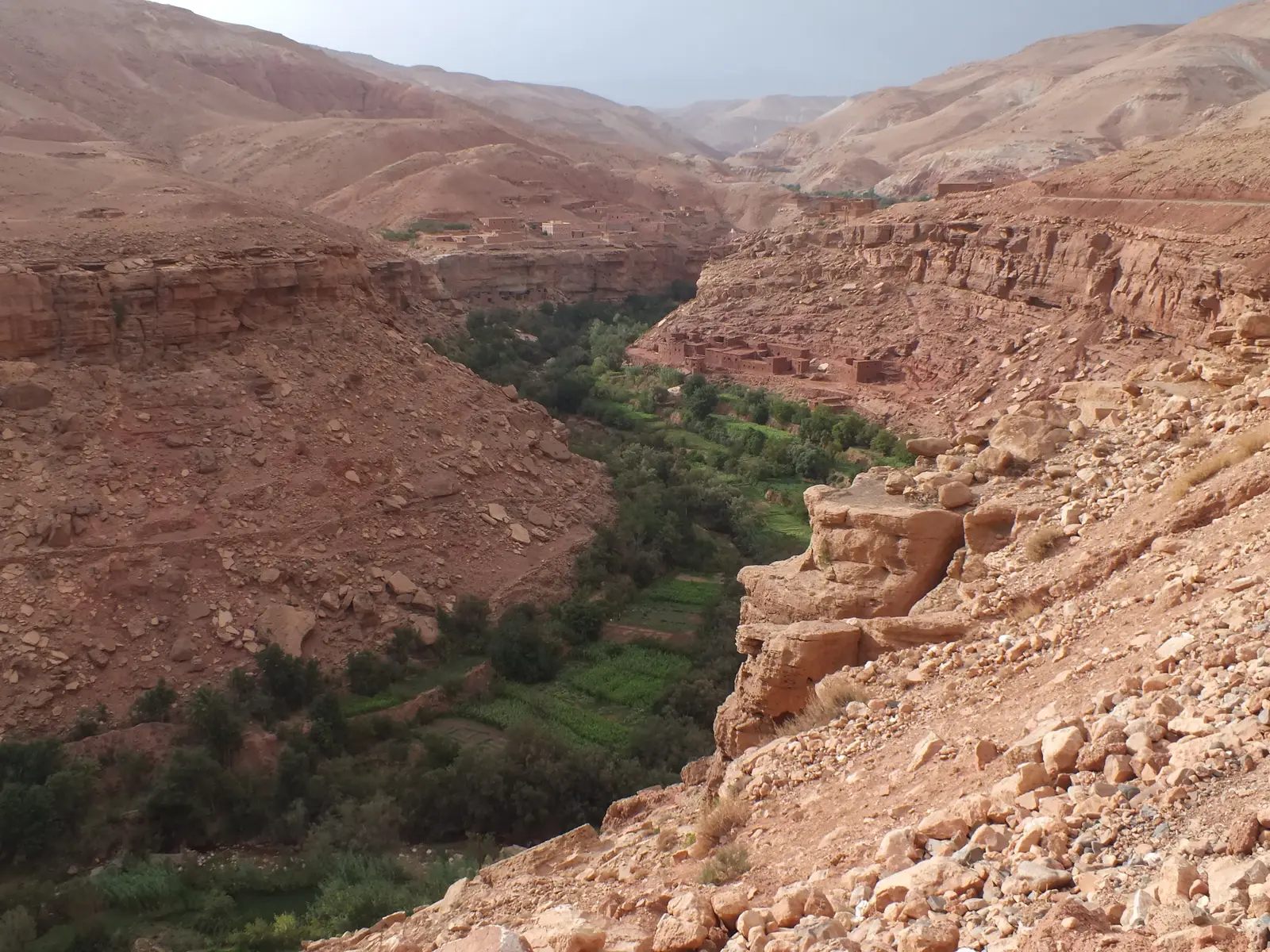 A deep red-rock gorge in the Ounila valley with a strip of green fields along the river at the bottom