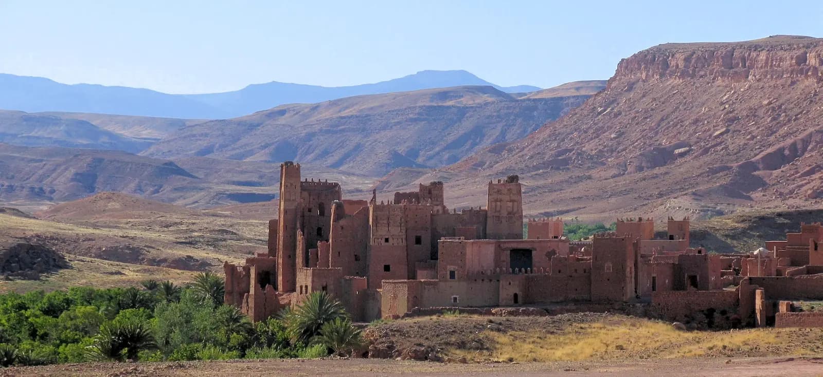 The towers of Tamdaght Kasbah among palms, with bare hills and a flat-topped ridge behind