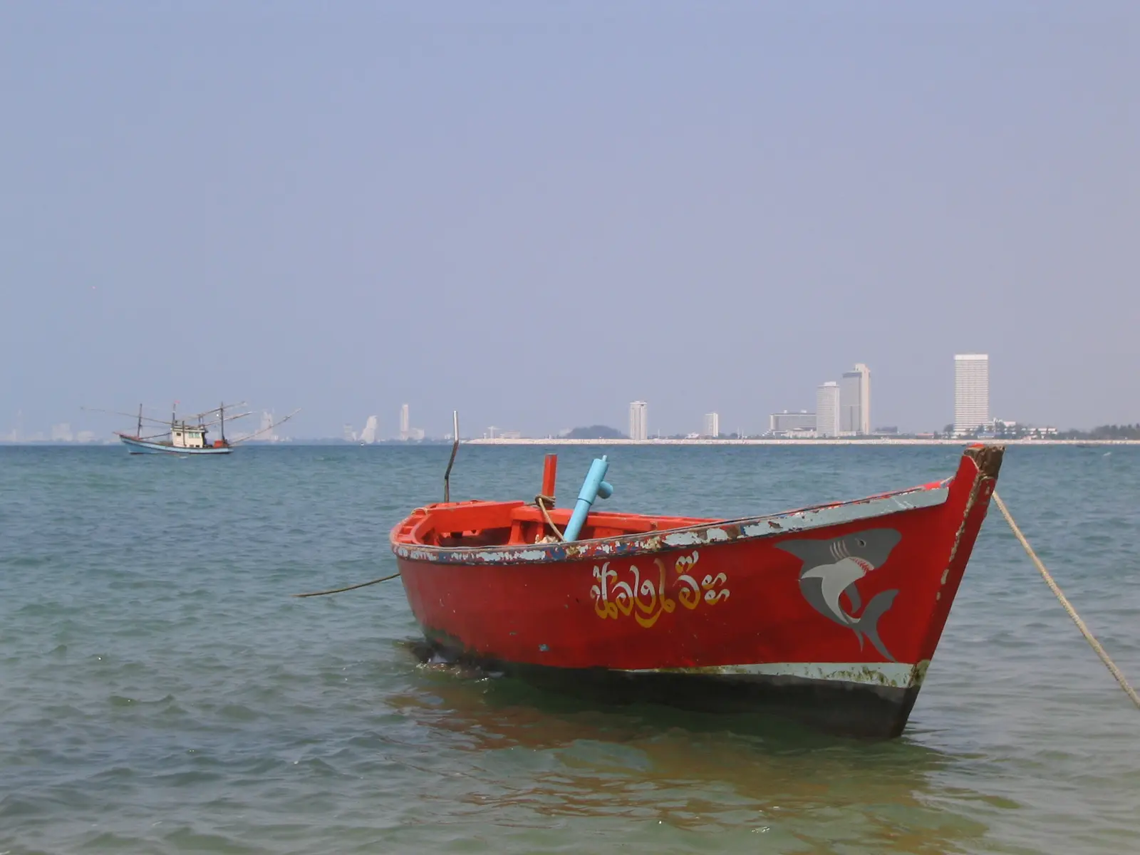 A red wooden fishing boat moored in shallow water at Bang Saray, with a trawler and distant high-rises behind