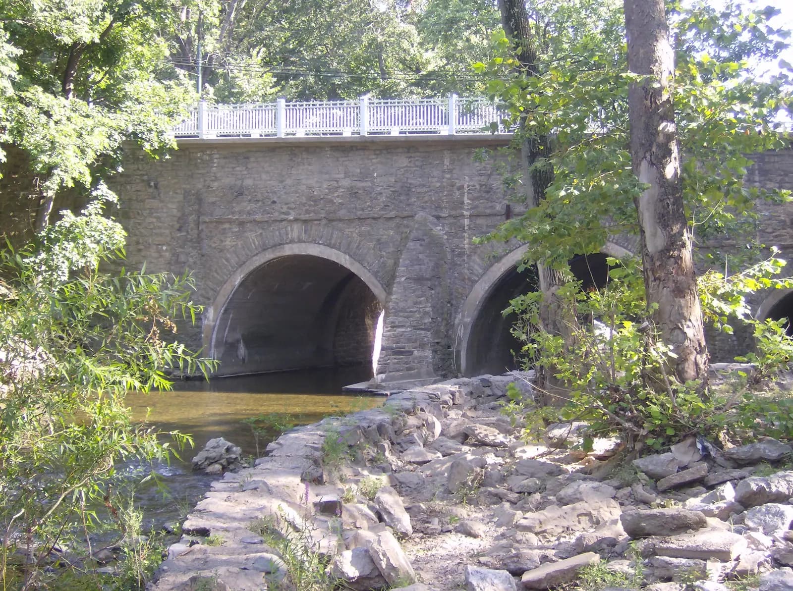 The stone arches of the Frankford Avenue Bridge over Pennypack Creek, seen from the rocky creek bank