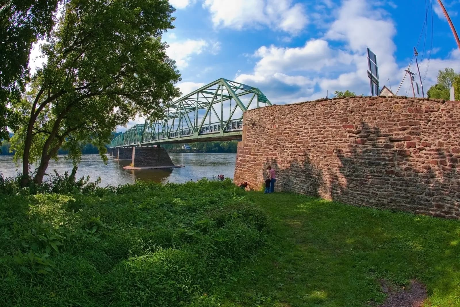 The green truss of the Uhlerstown–Frenchtown Bridge crossing the Delaware, seen from the grassy riverbank beside a stone wall