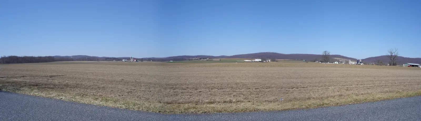 A wide, flat field in the Oley Valley under a blue sky, with farm buildings and low hills on the horizon