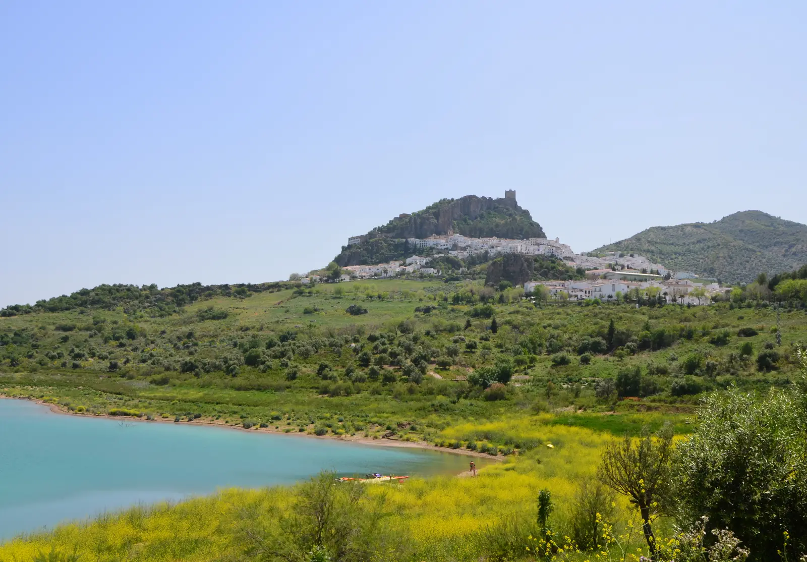 Zahara de la Sierra and its castle on a hill above the turquoise edge of the reservoir, with yellow wildflowers in front