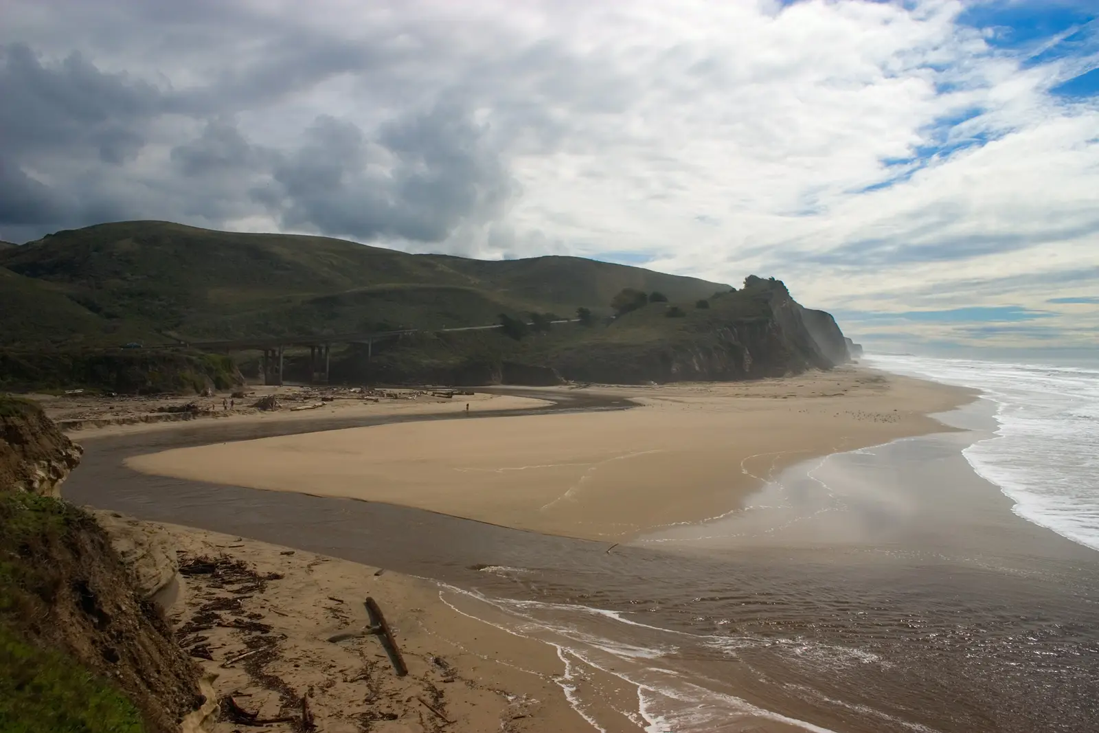 San Gregorio Creek running out across the beach below green bluffs, with the Highway 1 bridge behind