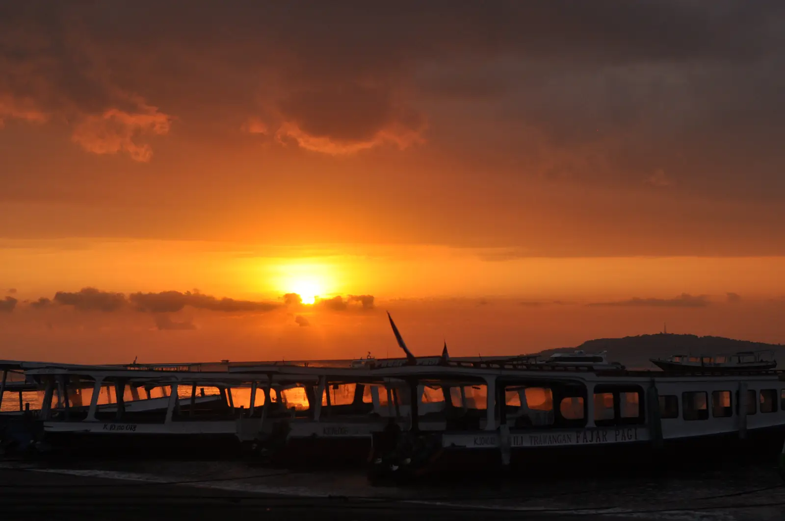 A row of moored wooden boats silhouetted against an orange sunset at Bangsal harbour