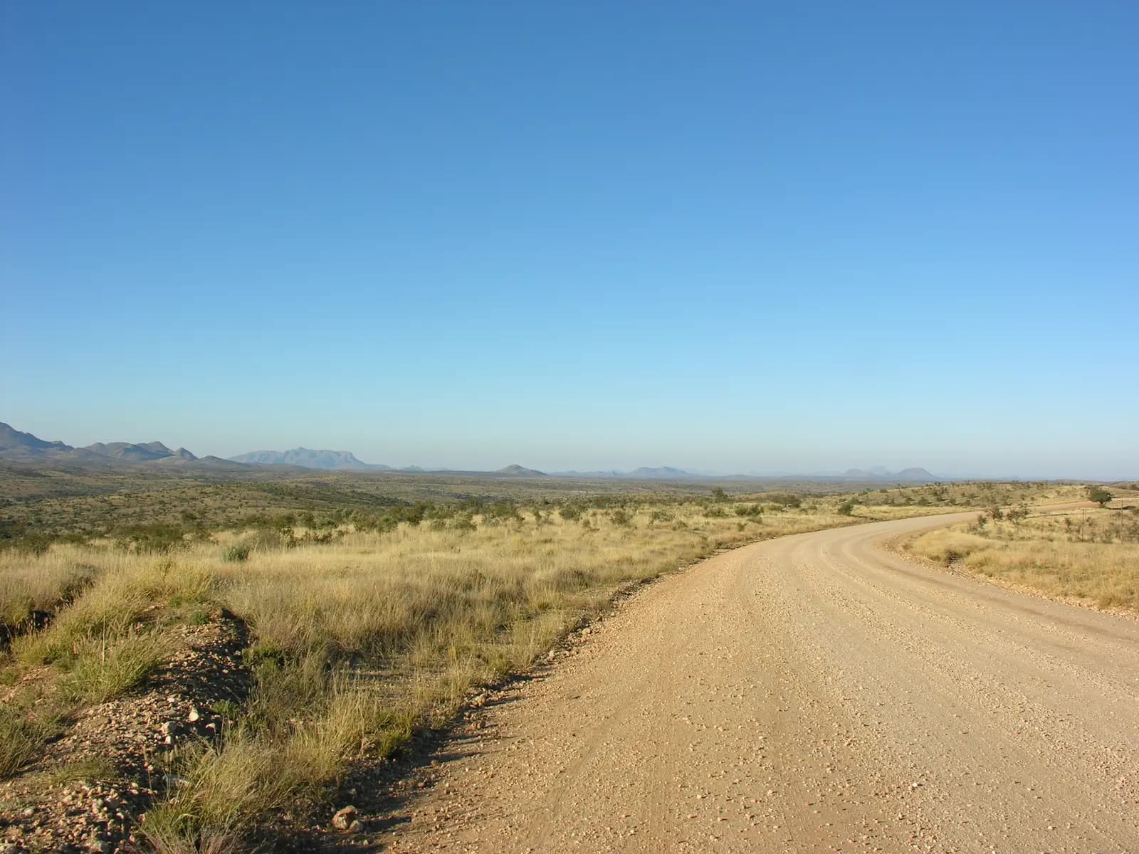 The gravel C26 bending through dry grass and thorn scrub, with distant hills on the horizon, near the top of the Kupferberg Pass